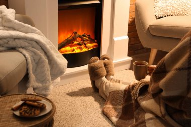 Terlikli bir kadın ve evinde şöminenin yanında oturan bir fincan çayla.