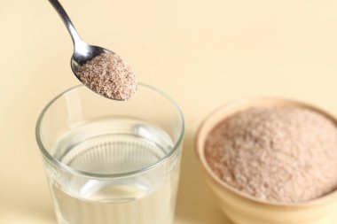 Bej arka planda psyllium kabuklu bir bardak su ve kaşık, yakın plan.