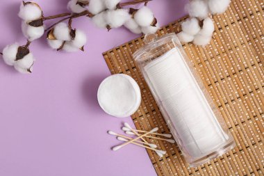 Kozmetik pedleri, bezleri, pamuk dalı ve leylak arkaplanda bambu paspası olan bir kap.