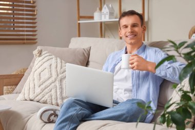 Bir fincan sıcak çayla kanepede oturan ve dizüstü bilgisayar kullanan genç adam.