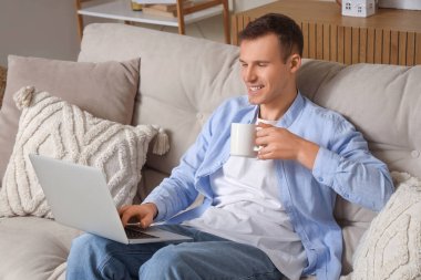 Kanepede oturmuş bir fincan sıcak çayla gülümseyen genç adam ve dizüstü bilgisayar.