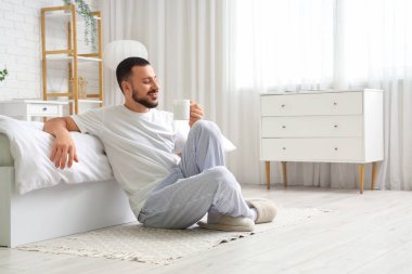 Yakışıklı genç adam yerde oturuyor ve yatak odasında çay içiyor.