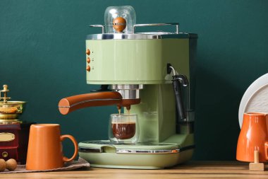 Mutfaktaki yeşil duvarın yanındaki ahşap masada bir bardak sıcak espresso ile modern kahve makinesi.