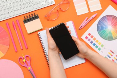 Turuncu arka planda boş cep telefonu ve kırtasiyesi olan kadın grafik tasarımcısı, üst görünüm