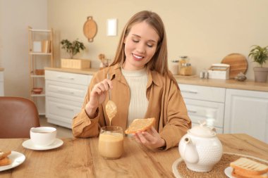 Genç bir kadın mutfakta kaşıkla fıstık ezmesini tostun üzerine sürüyor.