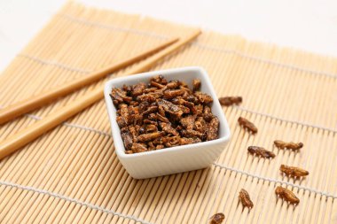Kızarmış böcekler ve beyaz arka planda bambu hasırı üzerinde yemek çubuklarıyla kase.
