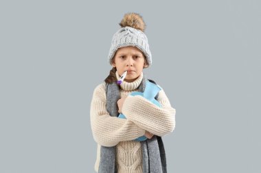 Gri arka planda termometresi ve sıcak su torbası olan hasta küçük kız.