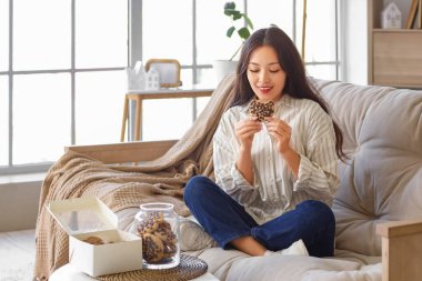 Genç Asyalı kadın evdeki kanepede kurabiye yiyor.