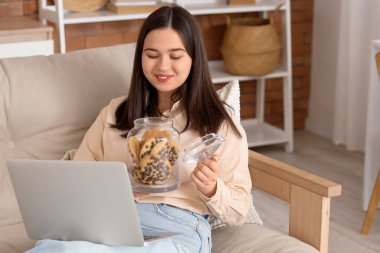 Elinde kurabiye kavanozu ve dizüstü bilgisayarı olan genç bir kadın evdeki kanepede yatıyor.