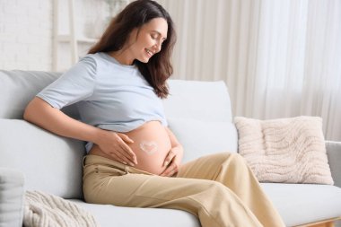 Kanepede oturan, göbeğinde kremden yapılmış kalbi olan hamile bir kadın.