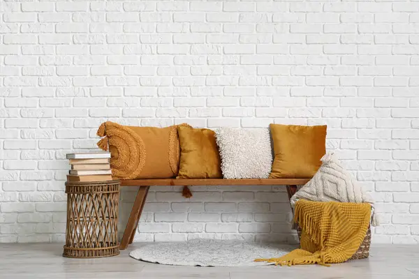 Dekoratif yastıklı ahşap bank, beyaz tuğla duvarın yanında masa ve sepet.