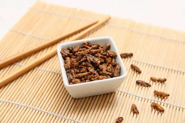 Kızarmış böcekler ve beyaz arka planda bambu hasırı üzerinde yemek çubuklarıyla kase.