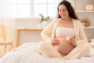 Genç hamile kadın yatak odasında vücut kremi sürüyor.