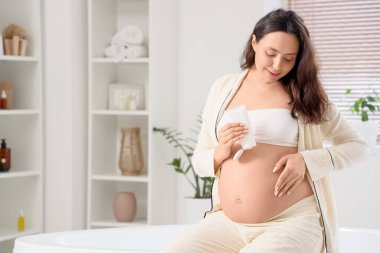 Genç hamile bir kadın banyoda vücut kremi sürüyor.