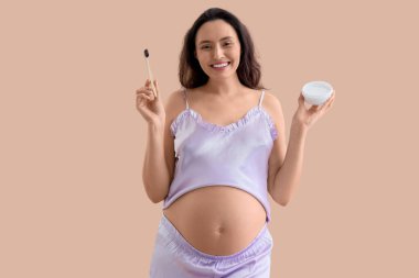 Bej arka planda diş fırçası ve pudrası olan hamile bir kadın.