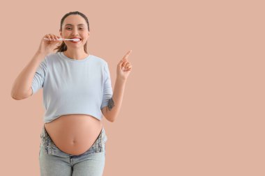 Hamile bir kadın dişlerini fırçalıyor ve bej arka planda bir şeye işaret ediyor.