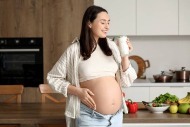 Mutfakta bir bardak yoğurt taşıyan hamile bir kadın.