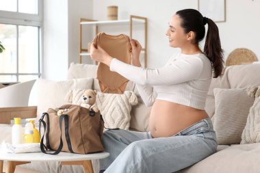 Bebek tulumu ve doğum çantası olan hamile bir kadın evdeki kanepede oturuyor.