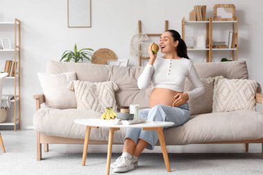 Genç hamile kadın evdeki kanepede elma yiyor.