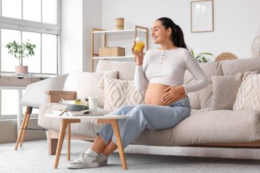 Genç hamile kadın evdeki kanepede meyve suyu içiyor.