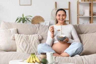 Genç hamile kadın evdeki kanepede müsli yiyor.