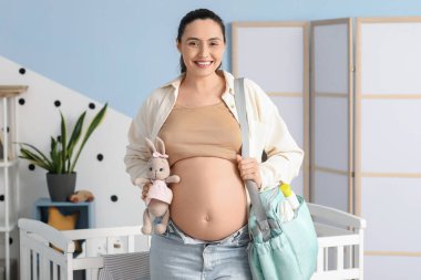 Çocuk odasında oyuncak ve doğum çantası taşıyan hamile bir kadın.