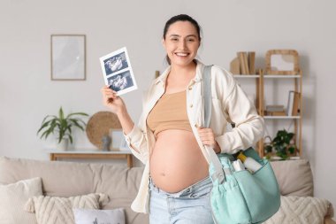 Evde sonogram görüntüsü ve doğum çantası olan hamile bir kadın var.