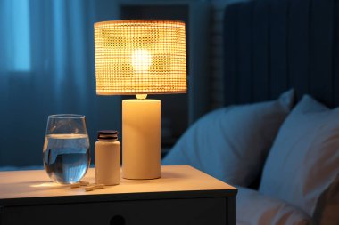 Geceleri yatak odasındaki masada bir bardak su ve parlayan lambayla birlikte melatonin hapları.