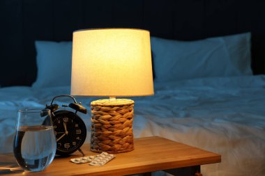 Çalar saat, melatonin hapları, bir bardak su ve geceleri yatak odasındaki masada parlayan lamba.