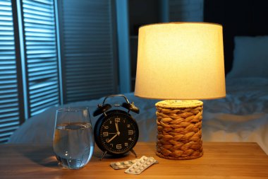 Çalar saat, melatonin hapları, bir bardak su ve geceleri yatak odasındaki masada parlayan lamba.