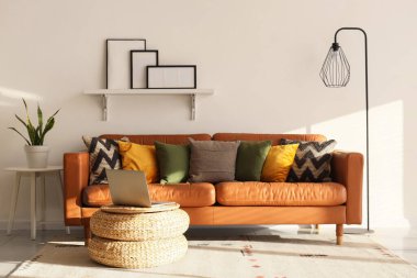 Oturma odasının içinde lamba, şık deri kanepe ve beyaz duvarın yanındaki hasır masada dizüstü bilgisayar.