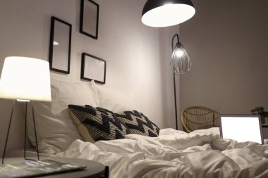 Modern dizüstü bilgisayarı olan rahat bir yatak ve gece şık yatak odasında parlayan lambalar.