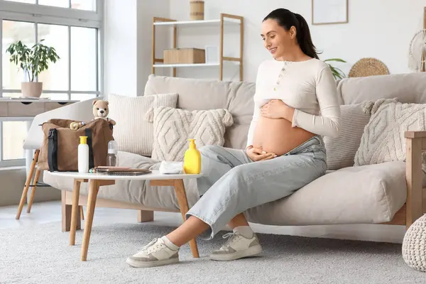 Doğum çantası ve bebek eşyaları olan hamile bir kadın evdeki kanepede oturuyor.