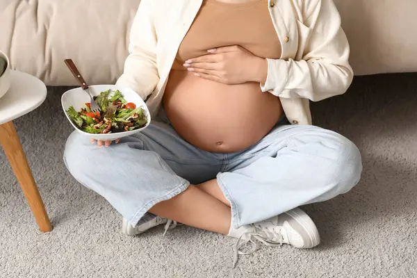 Evde halıya oturmuş sebze salatası yiyen hamile bir kadın.