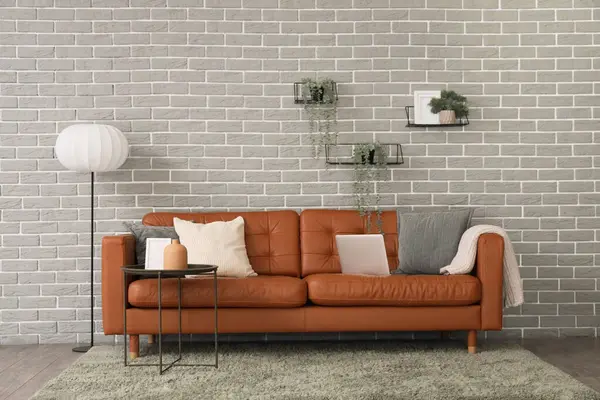 İçinde lamba, raflar, sehpa ve laptop olan oturma odasının içinde gri tuğlalı duvarın yanındaki şık deri koltukta.