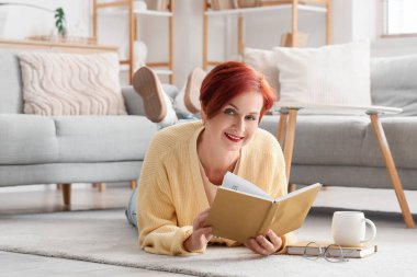 Güzel kızıl saçlı mutlu olgun kadın elinde kahve fincanıyla evde yerde kitap okuyor.