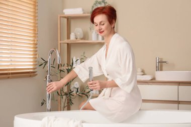 Güzel kızıl saçlı mutlu, bornozlu olgun bir kadın evde banyo yapmaya hazırlanıyor.