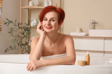 Güzel kızıl saçlı mutlu olgun kadın kozmetik ürünleriyle evde banyo yapıyor.