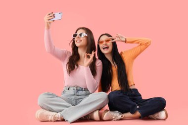 Mutlu genç kızlar pembe arka planda selfie çekiyorlar.