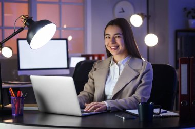 Akşam ofiste dizüstü bilgisayarla çalışan genç bir iş kadını.