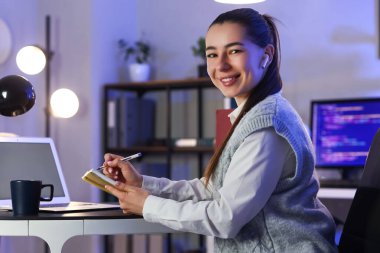 Kulaklıklı genç iş kadını, akşam ofiste deftere yazı yazıyor.