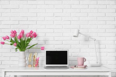 Modern dizüstü bilgisayarlı şık çalışma alanı, bir fincan çay ve beyaz tuğla duvarın yanında güzel lale çiçekleri.