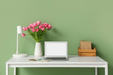 Modern dizüstü bilgisayarı, kitapları ve yeşil duvarın yanındaki güzel lale çiçekleriyle süslü çalışma alanı.
