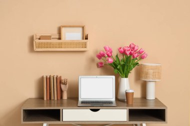 Modern dizüstü bilgisayar, kitaplar ve bej duvarın yanında güzel lale çiçekleri olan şık bir çalışma alanı.