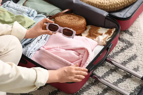 Genç bir kadın otel odasında giysileri ve güneş gözlükleriyle bavulunu boşaltıyor.