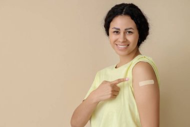 Genç Afro-Amerikalı kadın bej arka plandaki aşıdan sonra tıbbi yamaya işaret ediyor.