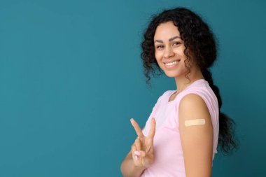 Uygulamalı tıbbi bandı olan genç bir Afro-Amerikalı kadın mavi arka planda aşıdan sonra zafer işareti gösteriyor.