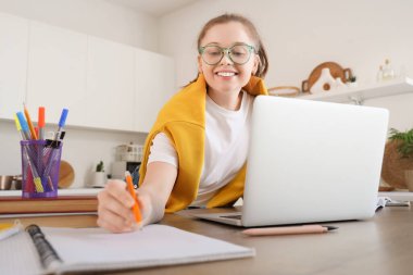 Not defteri ve dizüstü bilgisayarı olan genç kız mutfaktaki masada çalışıyor.