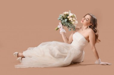Beyaz gelinlik içinde, çiçekli güzel gelin bej arka planda oturuyor.