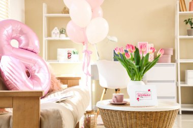 Vase with tulips, cup and greeting card for Women's Day on table in living room